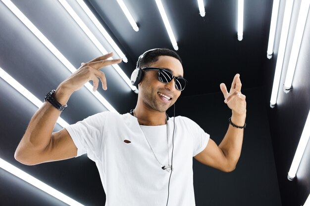 Young handsome smiling hipster black man in white outfit listening to music on headphones and dancing in hip hop style in disco night club, having fun