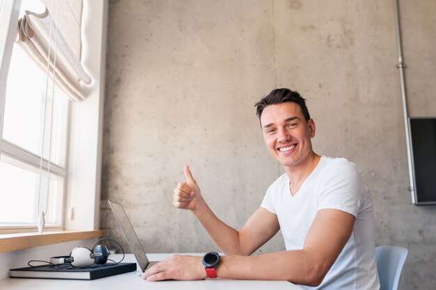 Young handsome smiling man in casual outfit sitting at table working on laptop, freelancer at home