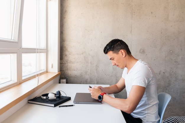 Young handsome smiling man in casual outfit sitting at table working on laptop staying at home alone, typing