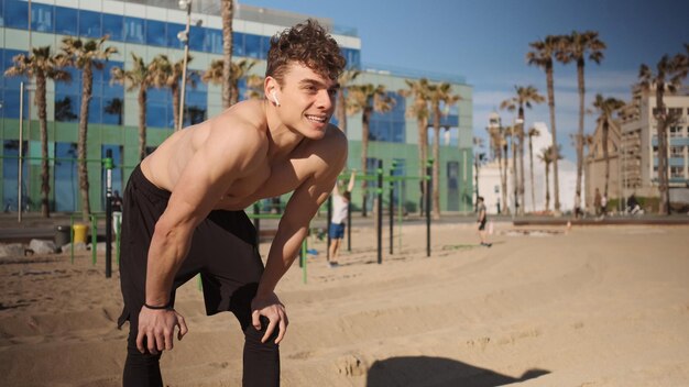 Young handsome smiling sporty man with wireless earphones resting during workout on city beach