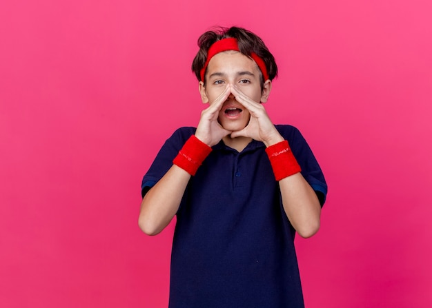 Young handsome sporty boy wearing headband and wristbands with dental braces looking at camera keeping hands around mouth calling out to someone isolated on crimson background with copy space