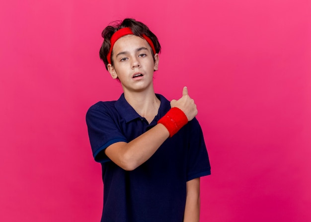 Young handsome sporty boy wearing headband and wristbands with dental braces looking at camera pointing behind isolated on crimson background with copy space