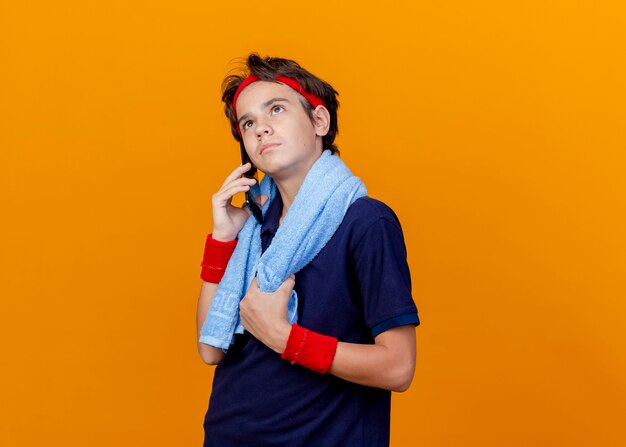 Young handsome sporty boy wearing headband and wristbands with dental braces and towel around neck grabbing towel talking on phone looking up isolated on orange background with copy space