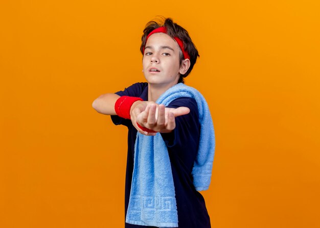 Young handsome sporty boy wearing headband and wristbands with dental braces and towel on shoulder standing in profile view isolated on orange wall