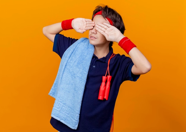 Young handsome sporty boy wearing headband and wristbands with dental braces and towel with jump rope on shoulders keeping hands on eyes isolated on orange wall