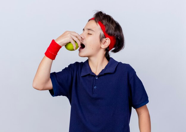 Young handsome sporty boy wearing headband and wristbands with dental braces turning head to side biting apple with closed eyes isolated on white background