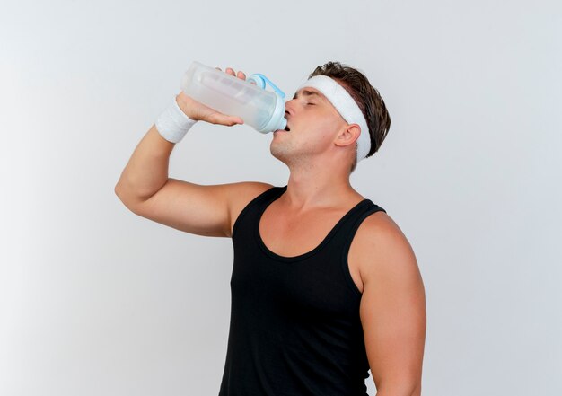 Young handsome sporty man wearing headband and wristbands drinking water from water bottle with closed eyes isolated on white background with copy space