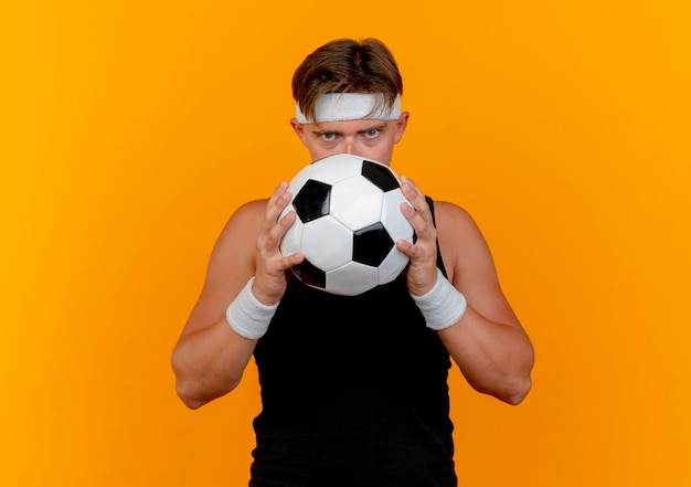 Молодой красивый спортивный мужчина с повязкой на голову и браслетами, держащими и смотрящими сзади на футбольный мяч на камеру, изолированную на оранжевом фоне