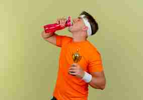 Free photo young handsome sporty man wearing headband and wristbands holding winner cup and drinking water from water bottle isolated on olive green background with copy space