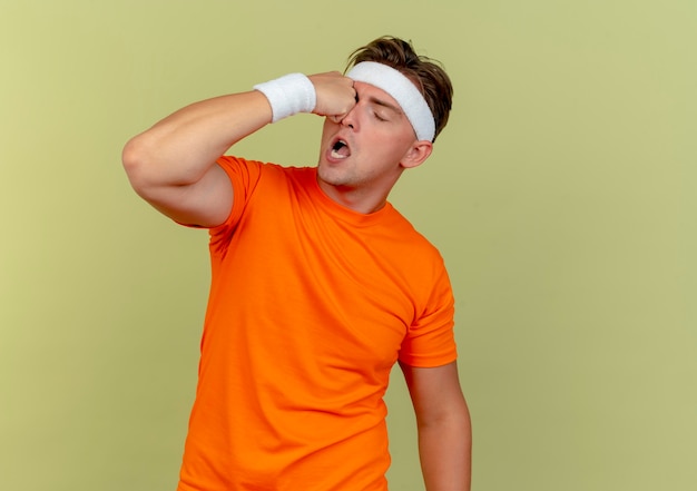 Young handsome sporty man wearing headband and wristbands kicking himself in eye isolated on olive green background with copy space