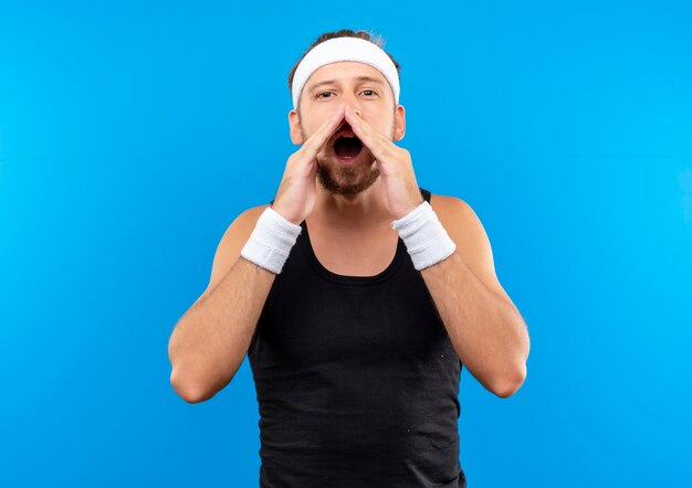 Young handsome sporty man wearing headband and wristbands putting hands around mouth calling someone isolated on blue space