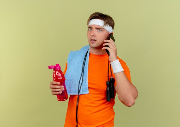 Young handsome sporty man wearing headband and wristbands with jump rope around neck and towel on shoulder holding water bottle and talking on phone isolated on olive green background with copy space