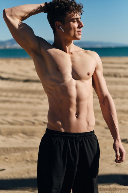 屋外でのトレーニングの後に休んでいる裸の胴体を持つ若いハンサムなスポーティな男。ビーチでポーズをとる魅力的なフィットネス男性モデル