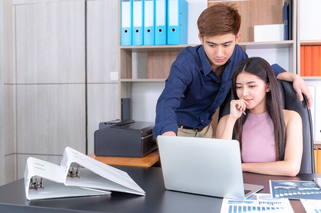 Young handsome and standing near pretty working woman sitting on chair at startup office 