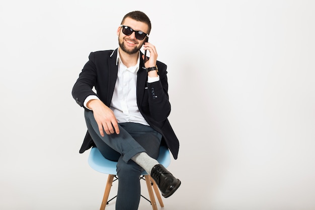 Giovane uomo bello alla moda hipster in giacca nera, stile di affari, camicia bianca, isolato, seduto rilassato sulla sedia da ufficio, parlando sullo smartphone, sorridente, felice, positivo, occhiali da sole