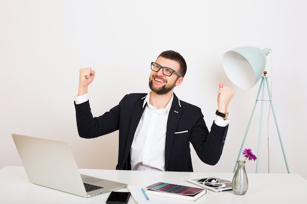 Young handsome stylish hipster man in black jacket sitting at office table, business style, white shirt, isolated, working on laptop, start up, work place, victory