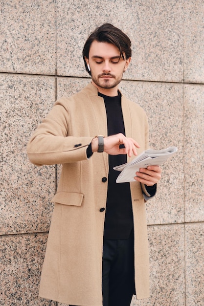 Young handsome stylish man in beige coat with newspaper watching time on wristwatch outdoor