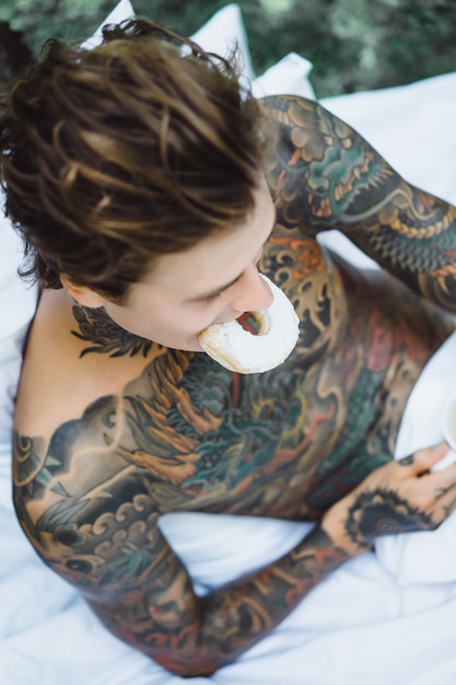 Foto gratuita giovane uomo bello tatuato facendo colazione a letto all'aperto all'aperto nel giardino