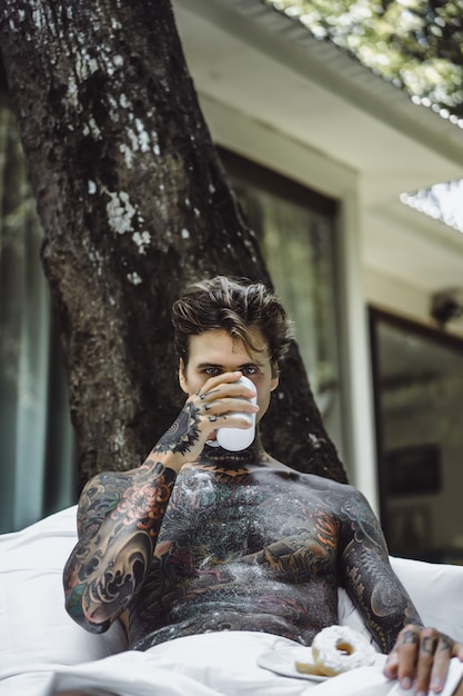 молодой красивый татуированный человек, завтрак в постели на открытом воздухе на открытом воздухе в саду