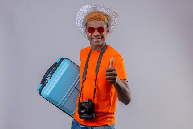 Young handsome traveler boy in summer hat wearing orange t-shirt holding travel suitcase smiling friendly showing thumbs up standing over white wall