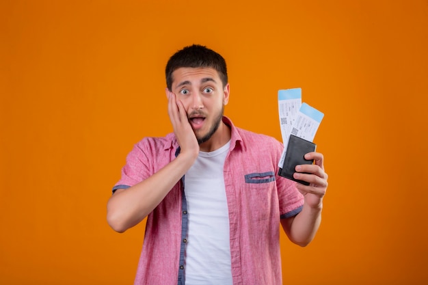 Young handsome traveler guy holding air tickets looking at camera surprised and amazed touching his face with arm standing over orange background