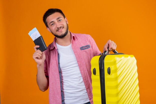 Young handsome traveler guy holding air tickets and suitcase looking at camera with confident smile positive and happy standing over orange background
