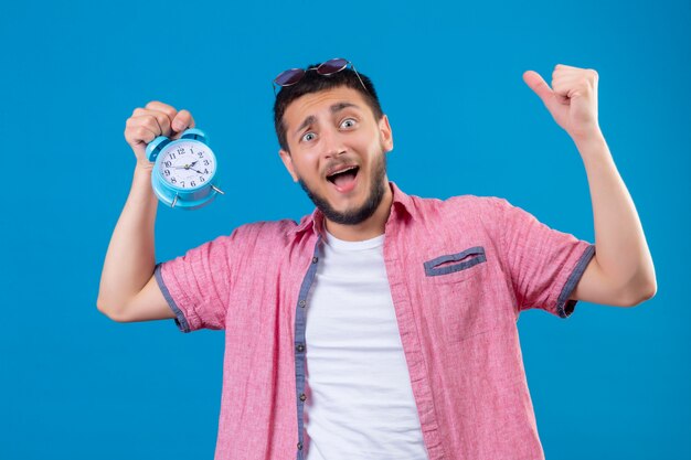 Young handsome traveler guy holding alarm clock looking surprised and amazed standing with raised hands over blue background