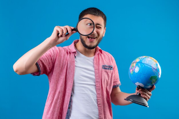 Young handsome traveler guy holding globe and looking at camera through magnifying glass smiling cheerfully standing over blue background