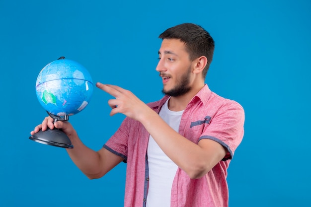 Молодой красивый путешественник парень держит глобус, глядя на него, улыбаясь со счастливым лицом, стоящим на синем фоне