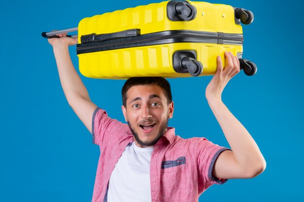 Young handsome traveler guy holding suitcase on his head looking joyful with happy smile on face standing over blue background