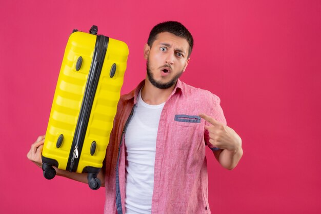 ピンクの背景の上に立っている彼自身を驚かせ、驚いて探しているスーツケースを持って若いハンサムな旅行者男