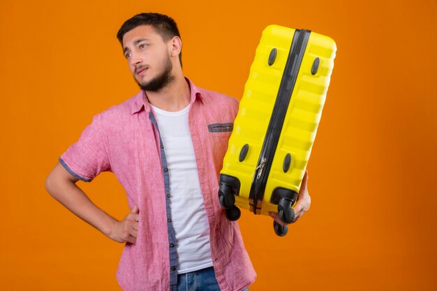 Young handsome traveler guy holding suitcase looking aside bored and tired with sad expression on face standing over orange background