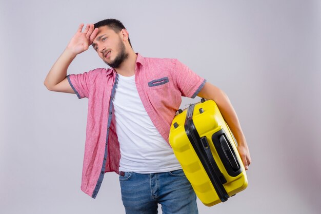 Молодой красивый путешественник парень держит чемодан, глядя в сторону, скучающий и усталый с грустным выражением лица, стоящего на белом фоне