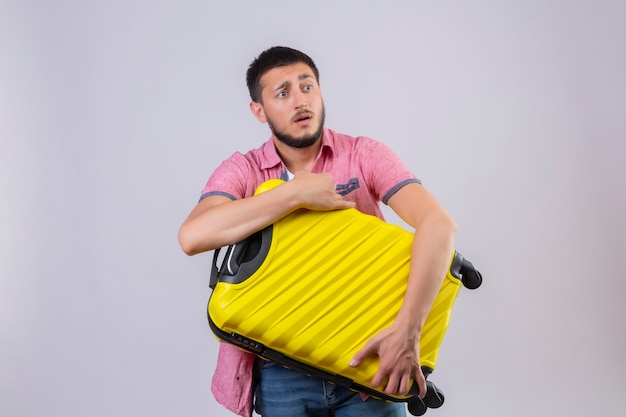 Young handsome traveler guy holding suitcase looking aside confused  with sad expression on face standing over white background