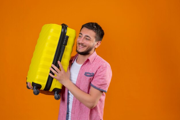 Young handsome traveler guy holding suitcase looking aside positive and happy smiling standing over orange background