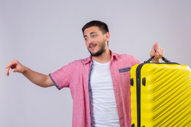 Young handsome traveler guy holding suitcase looking aside positive and happy smiling standing over white background
