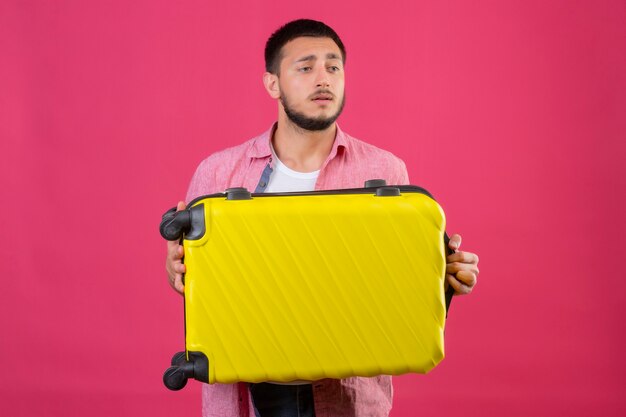 Young handsome traveler guy holding suitcase looking aside with sad expression on face standing over pink background
