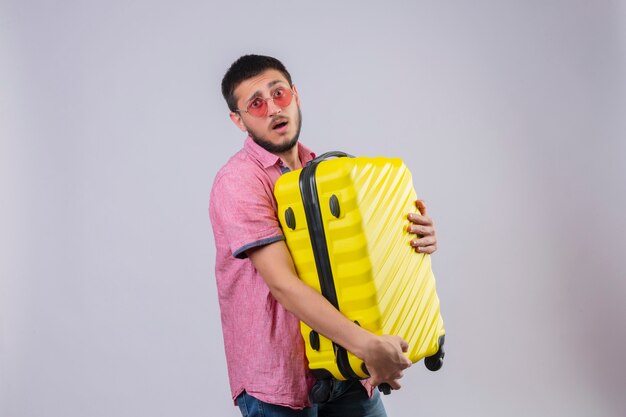 Young handsome traveler guy holding suitcase looking at camera confused with sad expression on face standing over white background