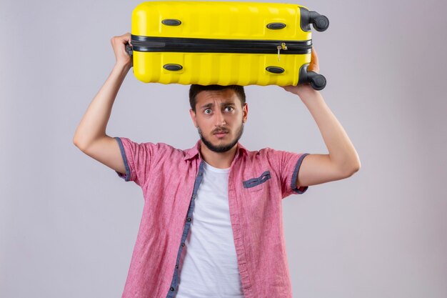 Young handsome traveler guy holding suitcase looking at camera confused  with sad expression on face standing over white background