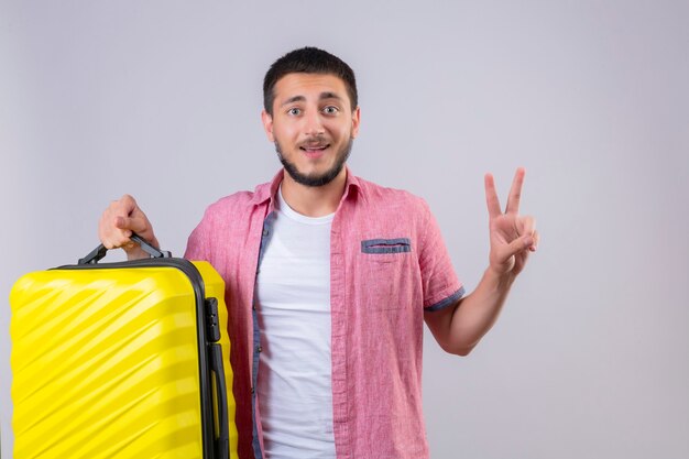 Young handsome traveler guy holding suitcase looking at camera smiling happy and positive showing number two or victory sign standing over white background