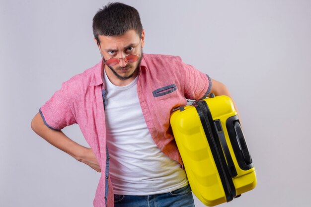 Young handsome traveler guy holding suitcase looking at camera with angry expression on face standing over white background