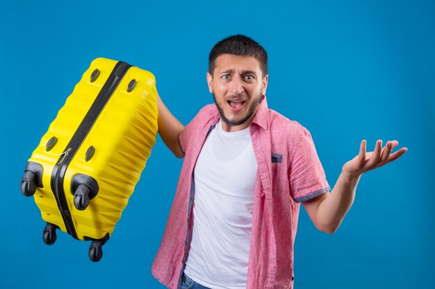 青い背景に失望して立っている探しているスーツケースを持って若いハンサムな旅行者男