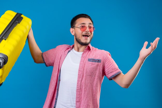 Young handsome traveler guy holding suitcase looking exited and happy standing with raised arms over blue background