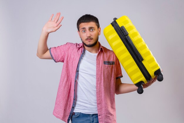 Young handsome traveler guy holding suitcase raising hand in surrender looking at camera with confuse expression on face standing over white background