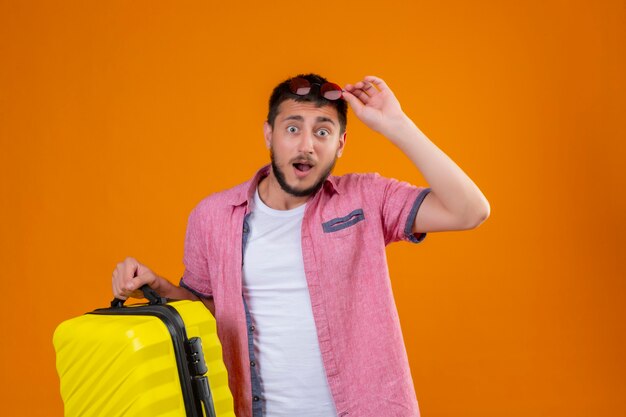 Young handsome traveler guy holding suitcase touching his sunglasses on head looking at camera surprised and exited standing over orange background