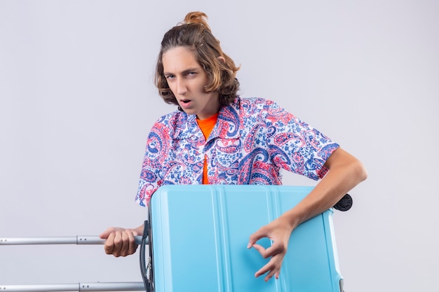 Young handsome traveler guy holding suitcase using as a guitar looking with serious confident expression on face standing over white background