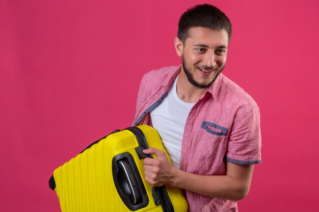 Молодой красивый путешественник с желтым чемоданом смотрит в сторону, весело улыбаясь со счастливым лицом, стоящим на розовом фоне