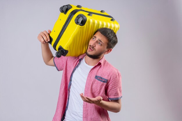 Молодой красивый путешественник, держащий желтый чемодан, выглядит смущенным, стоя на белом фоне