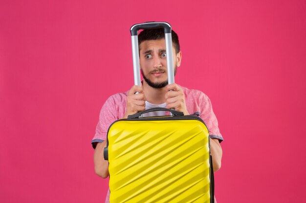 Young handsome traveler guy holding yellow suitcase looking confused with fear expression standing over pink background