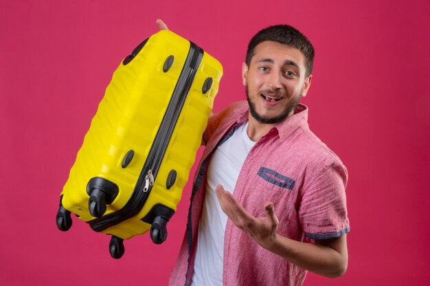 Молодой красивый путешественник, держащий желтый чемодан, весело улыбаясь, указывая рукой, стоящей на розовом фоне